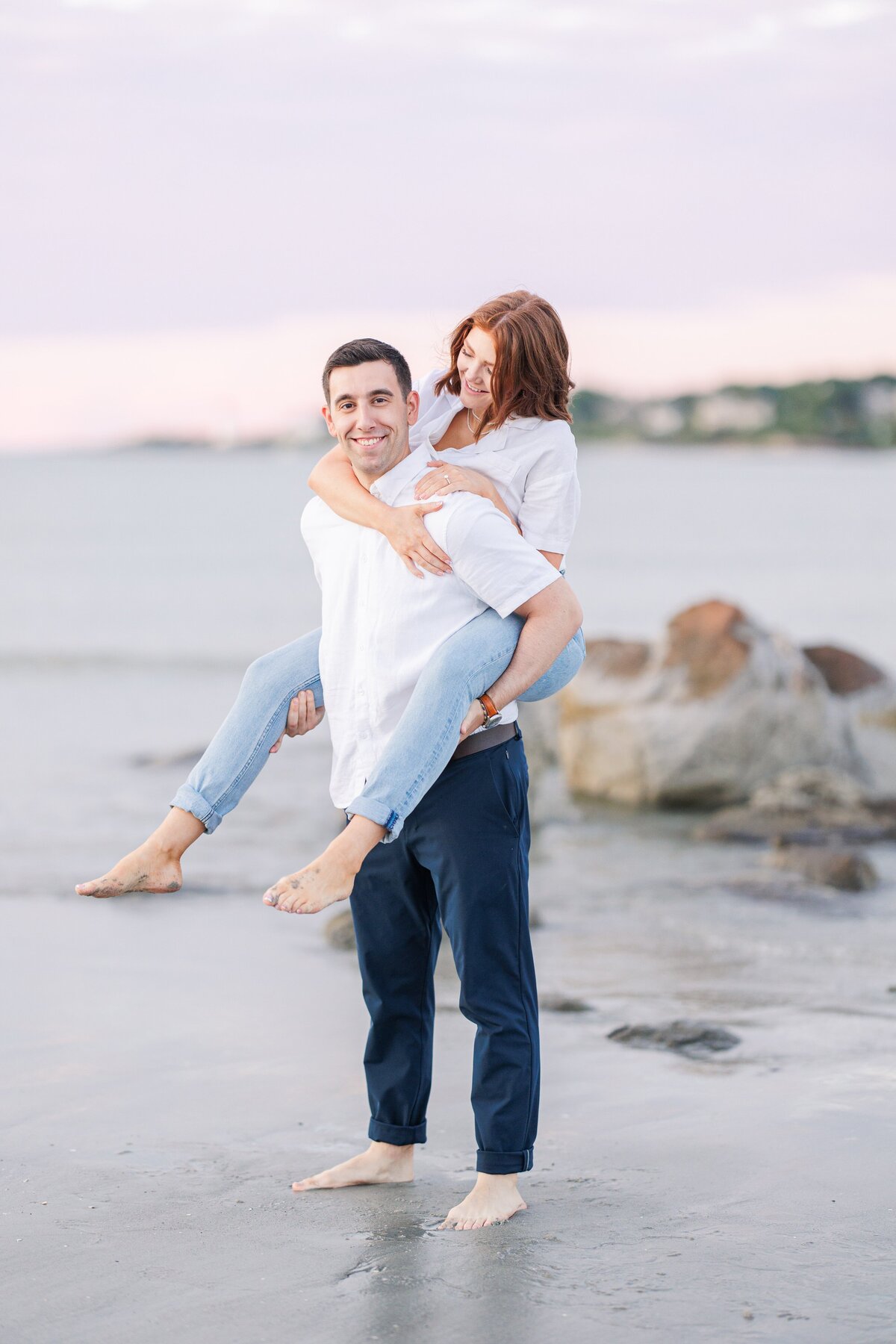 A woman smiling down at her fiance while he gives her a piggyback ride at their Wingaersheek Beach Gloucester engagement session.
