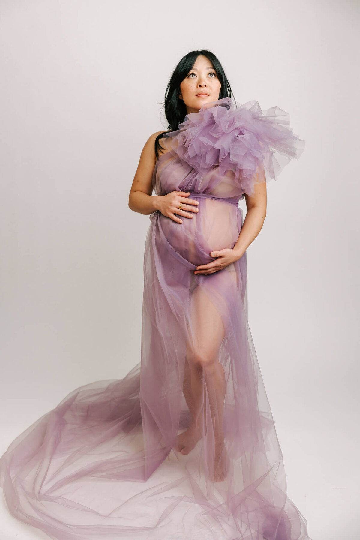 portrait of a mom wearing sheer purple dress with poof on sleeve. She had her hands on her belly and is looking up athe the light.