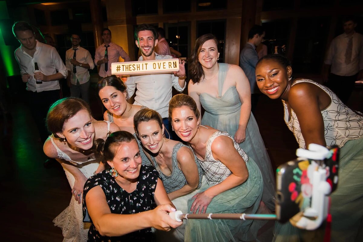 A group of bridesmaids and guests huddle around a photo booth sign at a wedding reception