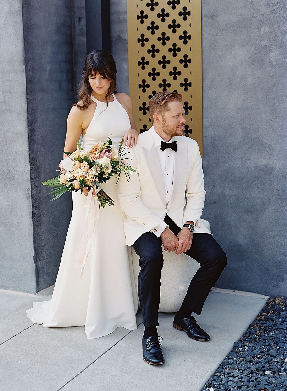 The bride, wearing a silk halter top sheath wedding dress and veil, holding a horizontal cascade bouquet of palm fronds and tropical flowers, as she stands behind the groom holding onto his shoulder in the courtyard of Clementine Hall. The groom, wearing a tuxedo with a white jacket, sits on a white stool in front of a gold filigree accent window against a gray wall.