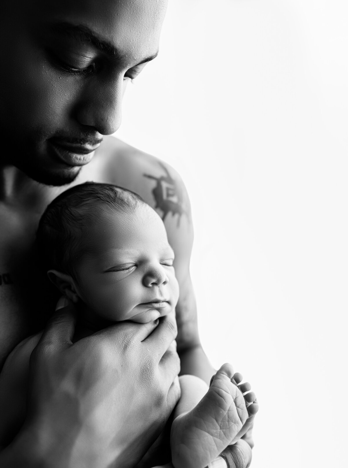 Black and white portrait of new father holding newborn son skin to skin contact while looking down at him