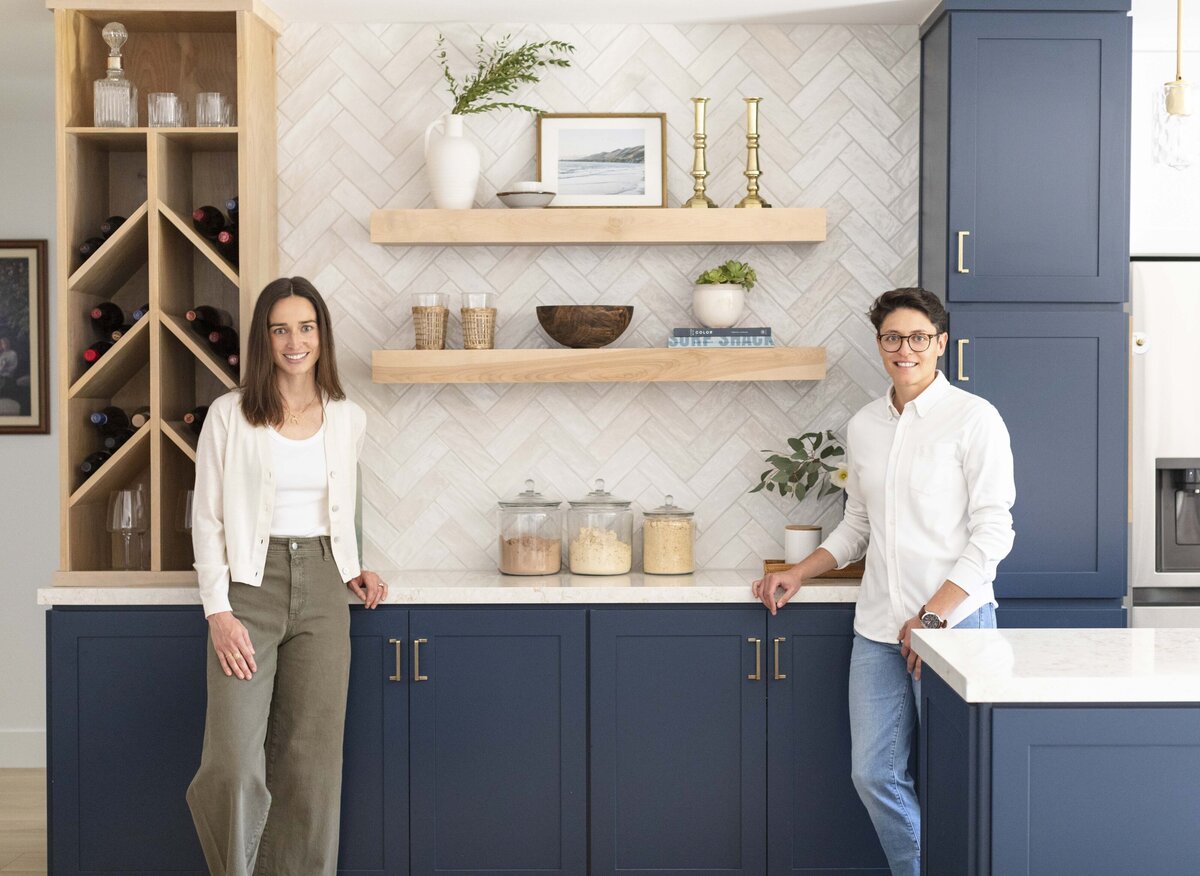 Two interior designers stand in a bright, modern kitchen featuring navy cabinetry, open wood shelving, and a wine rack, showcasing a beautifully styled space with warm coastal details, located in a Camarillo California home.