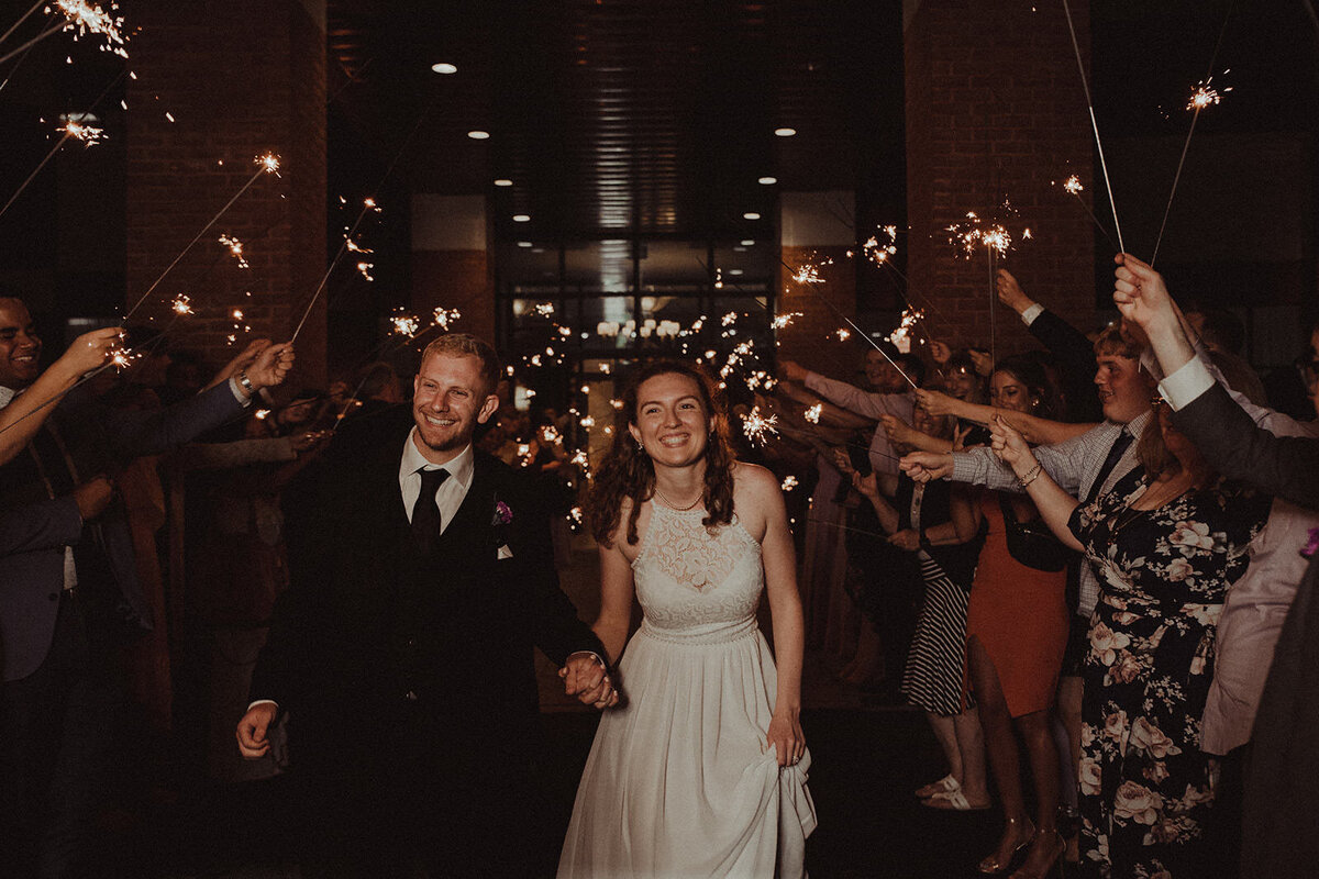 A couple holding hands as they are surrounded by their wedding guests holding sparklers 