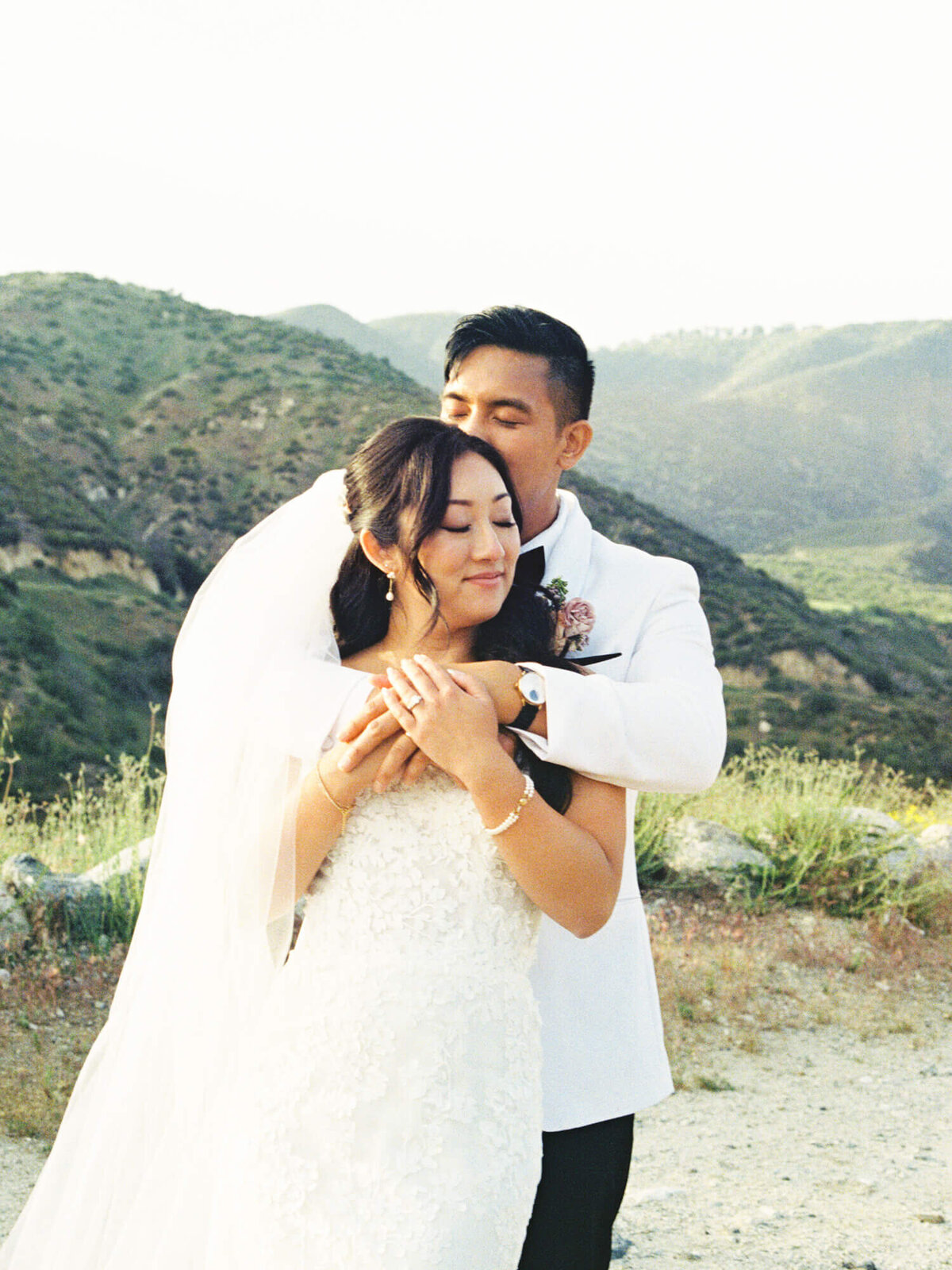 A bride and groom embrace lovingly against a scenic mountain backdrop. The bride wears a white lace dress and veil, evoking a serene, joyful atmosphere.