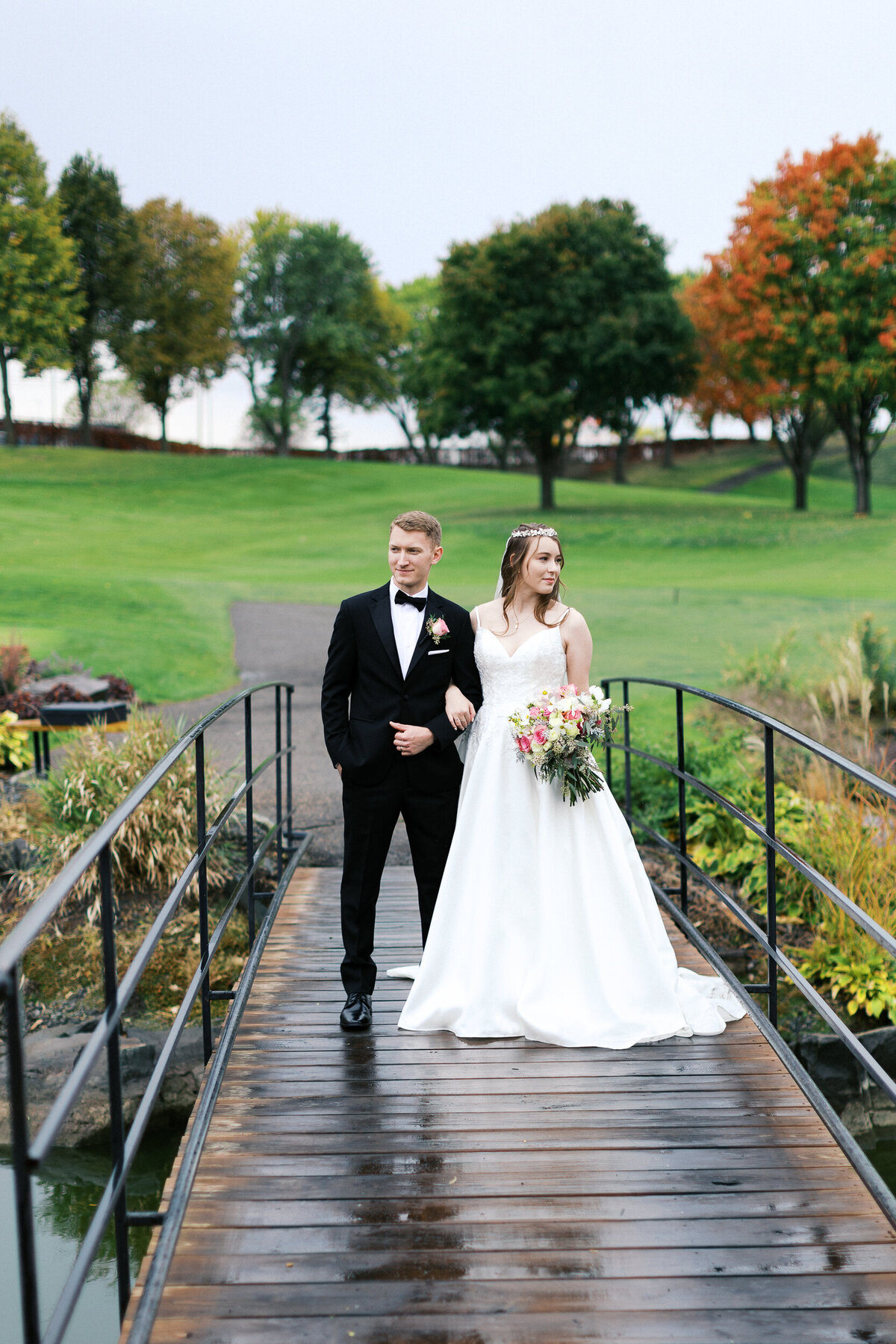 Newlyweds stand quietly under light rain on a bridge, facing opposite directions with arms linked — captured by Minnesota wedding photographer.