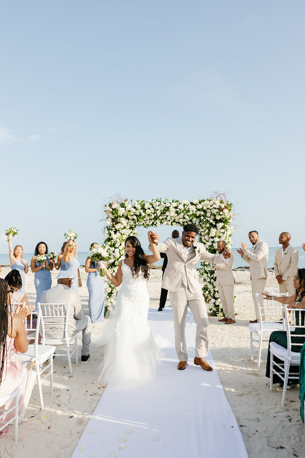 Newlyweds Andressa and Jonathan celebrate their wedding, surrounded by cheering guests and a stunning floral arch by the sea at Table Key, Islamorada. Captured by Claudia Amalia Wedding Photography.