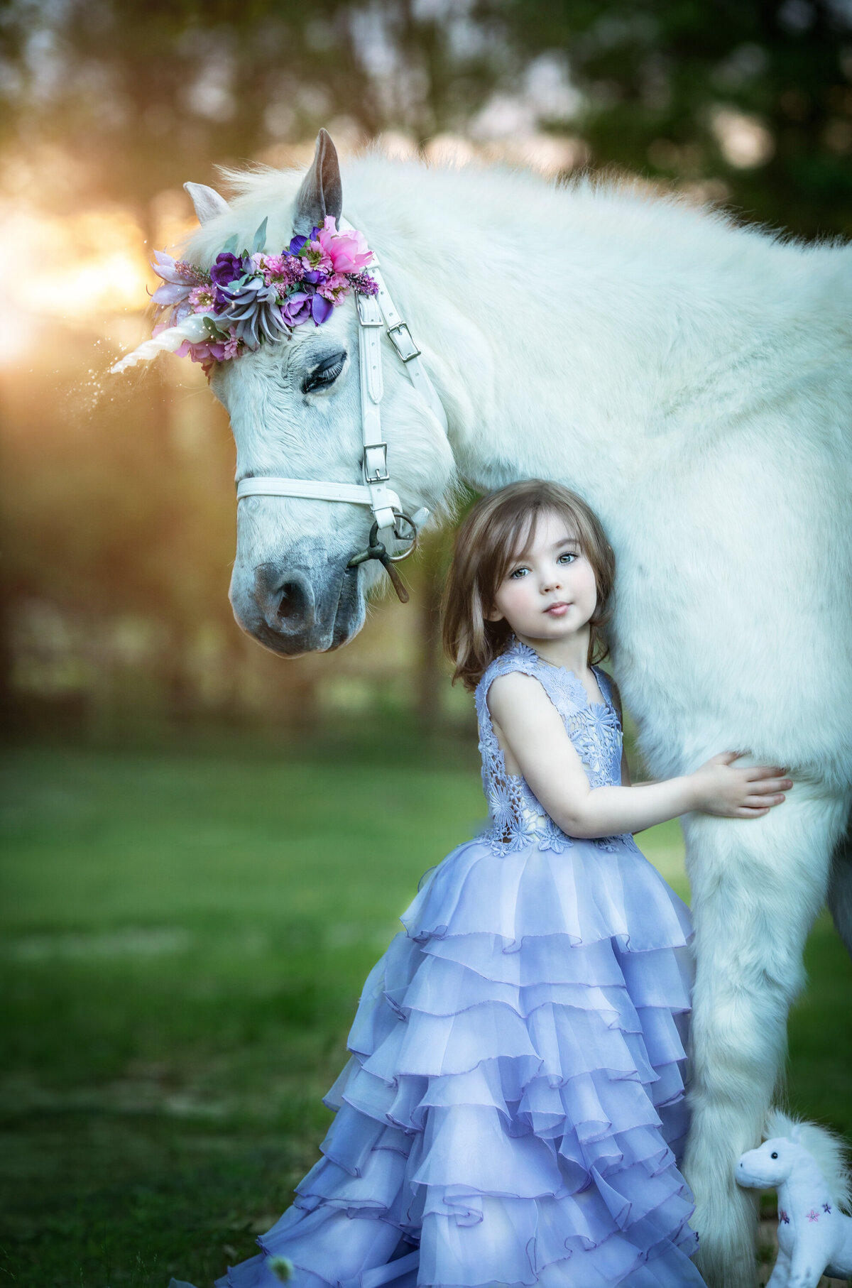 Girl in periwinkle dress by Butterfly Closet with shoulder length brown hair.  She is hugging a Welsh pony with a unicorn horn.  The horse is white.  She is holding onto its leg and there is a stuffed unicorn on the bottom right of the image.