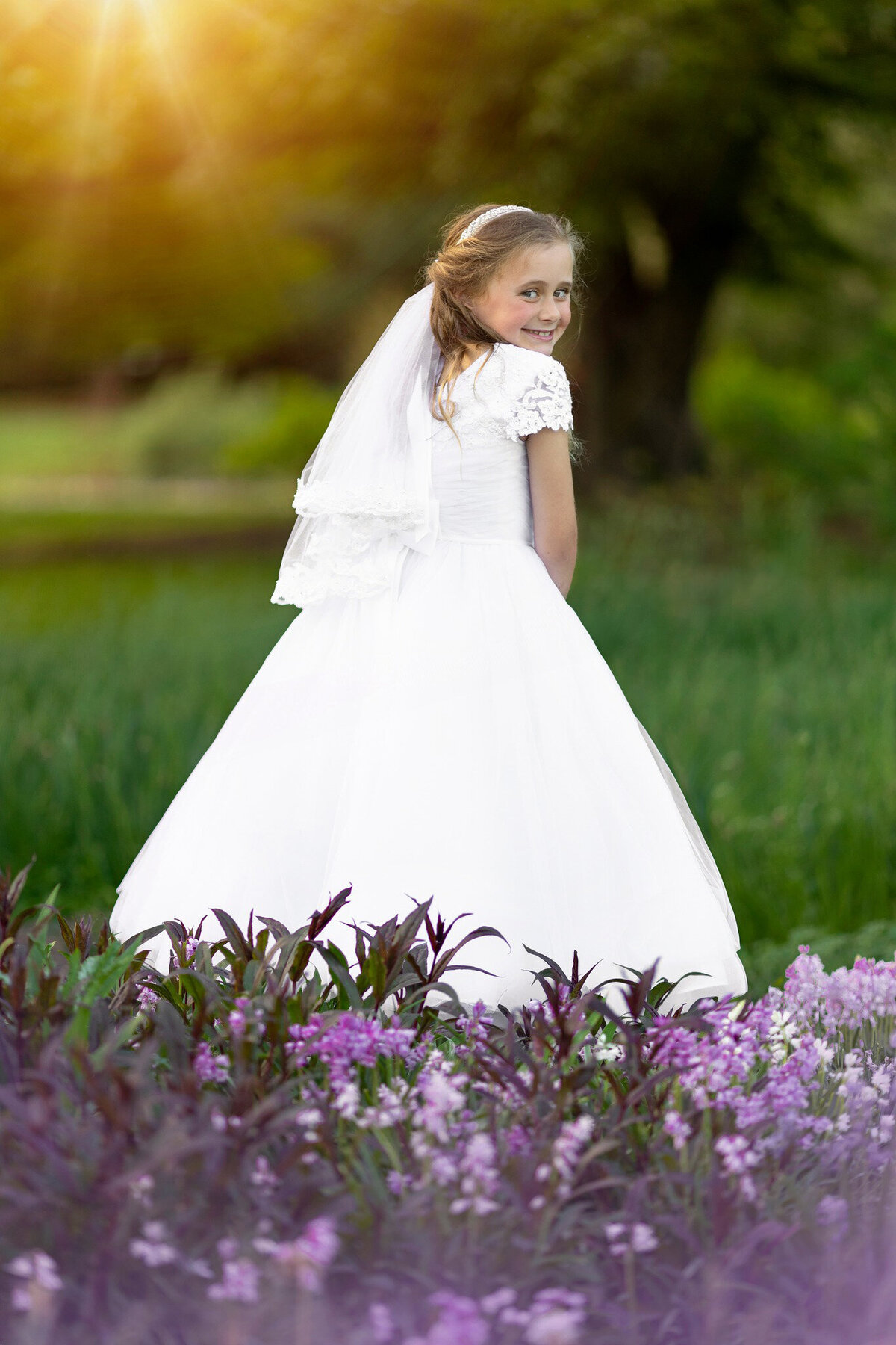 A young girl in a long white dress stands giggling over her shoulder in a garden with purple flowers for a New Jersey Communion Portrait Photographer