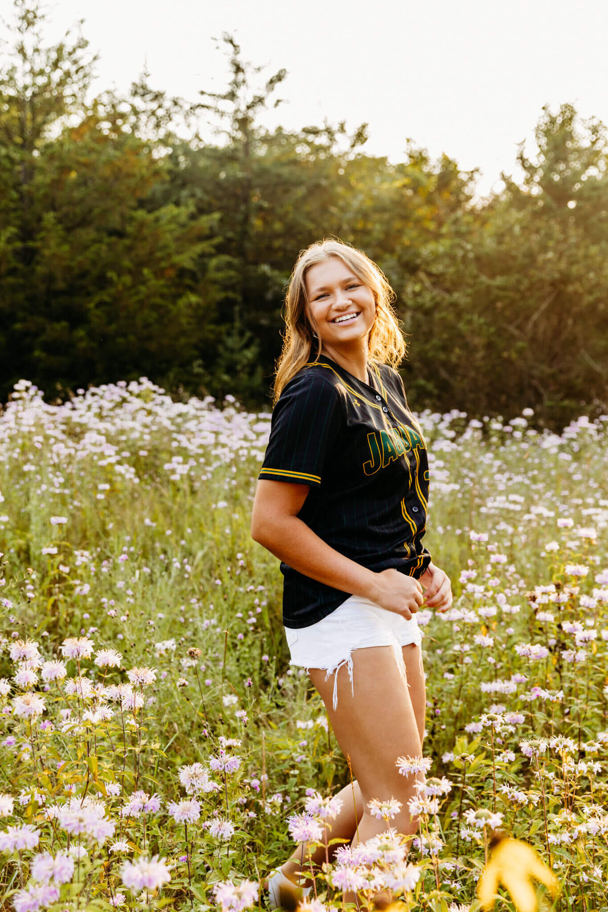 gorgeous teenage girl in a softball jersey and white shorts laughing as she plays in a field loaded with flowers