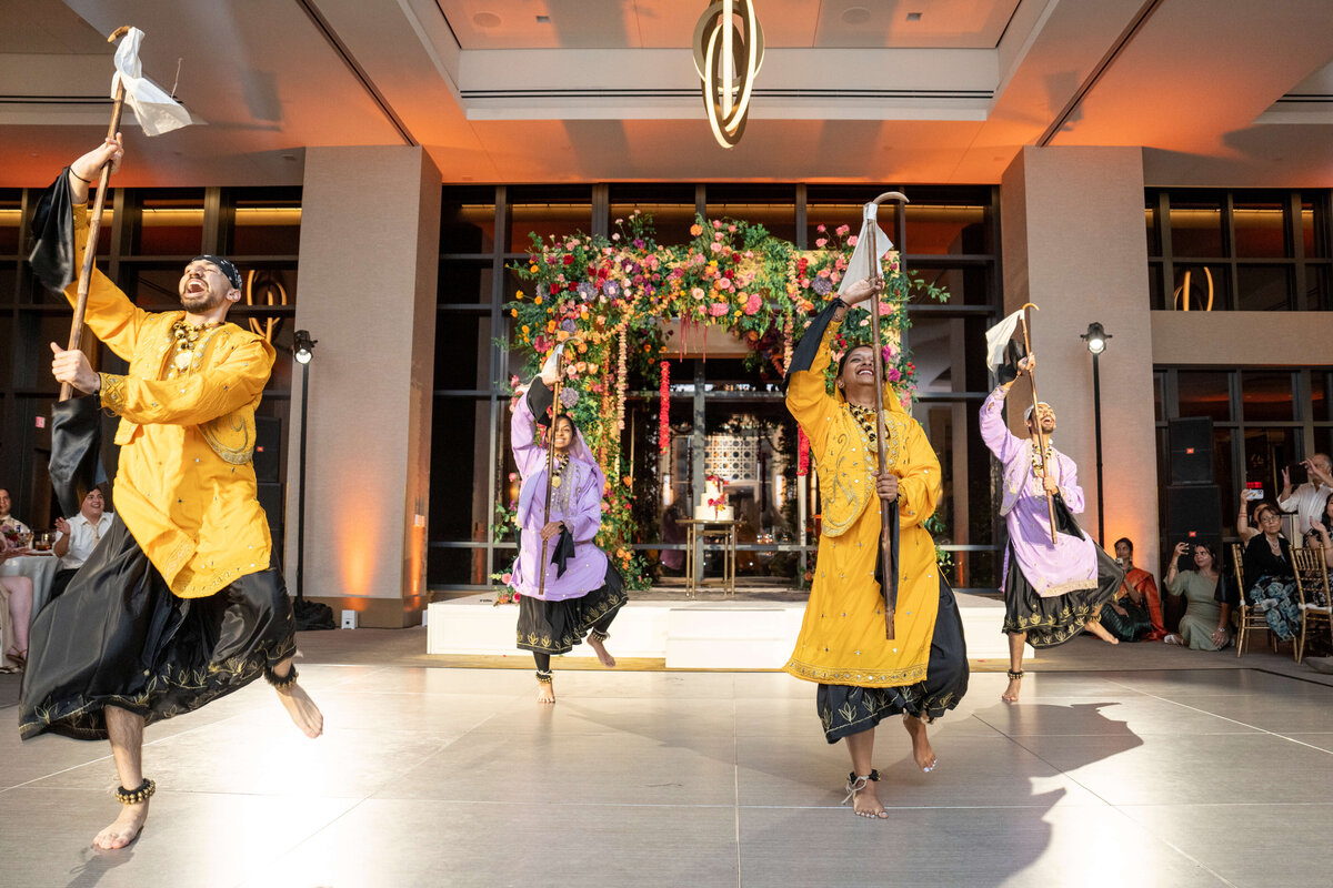 Four dancers in colorful traditional outfits perform joyfully with sticks in a bright, decorated venue. A floral backdrop and onlookers are visible in the background.