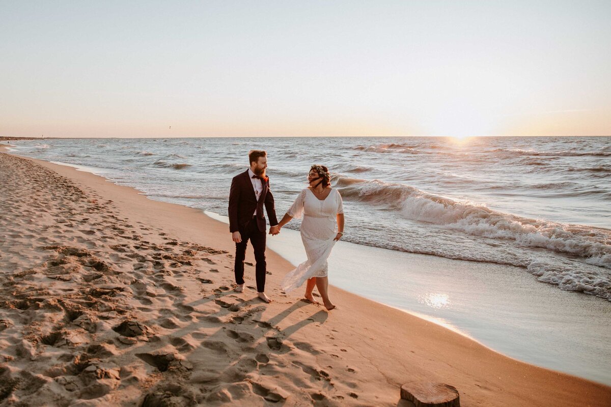 Bride and groom walking along Lake Huron shoreline at sunset. The bride is leading the groom, looking back at him. Warm golden light surrounds them.