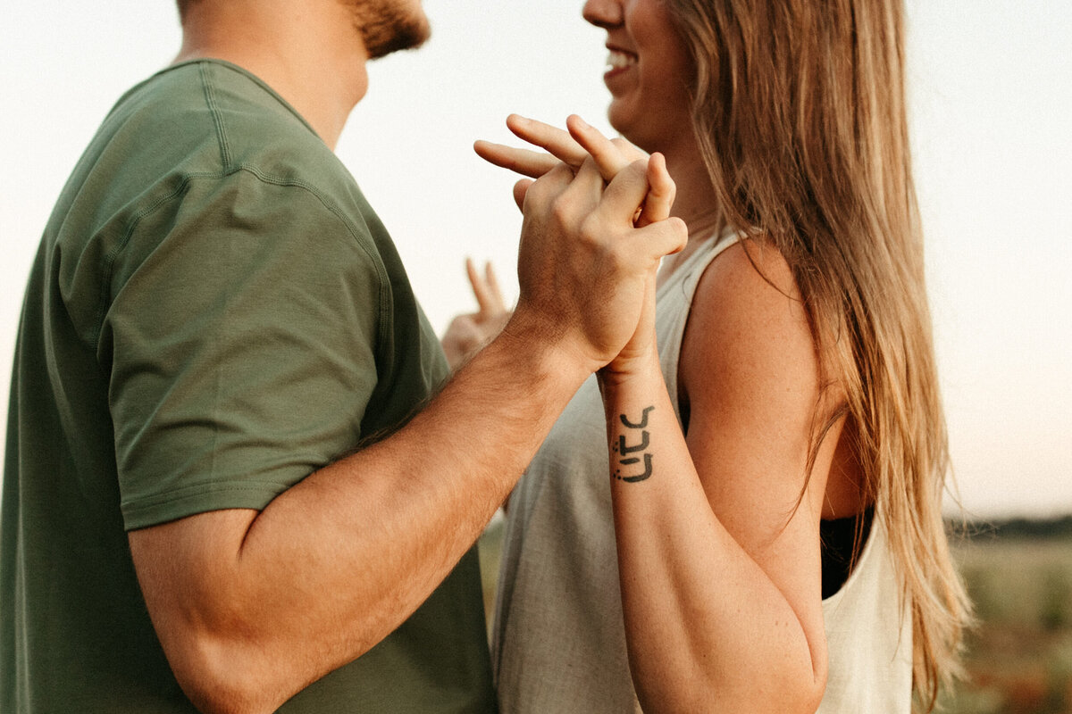 A guy and a girl are standing facing each other and intertwining their fingers as they smile at each other.