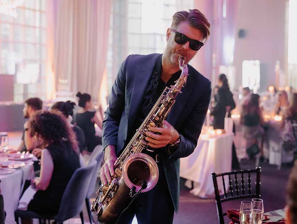 Live saxophone player performing at a formal indoor wedding with guests dining in the background.
