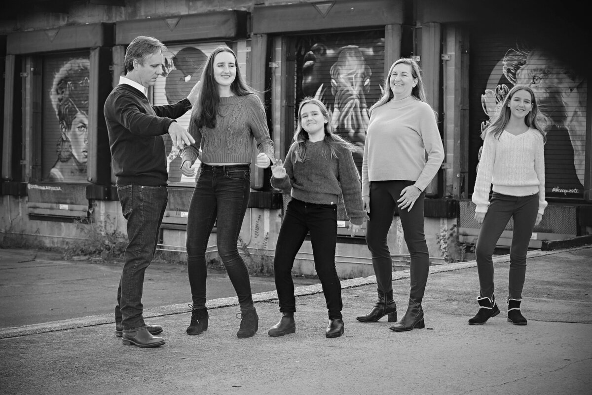 Playful family of five laughing and striking silly poses on ramp in front of colorful mural doors at Camp North End Charlotte NC. Photo in black and white.