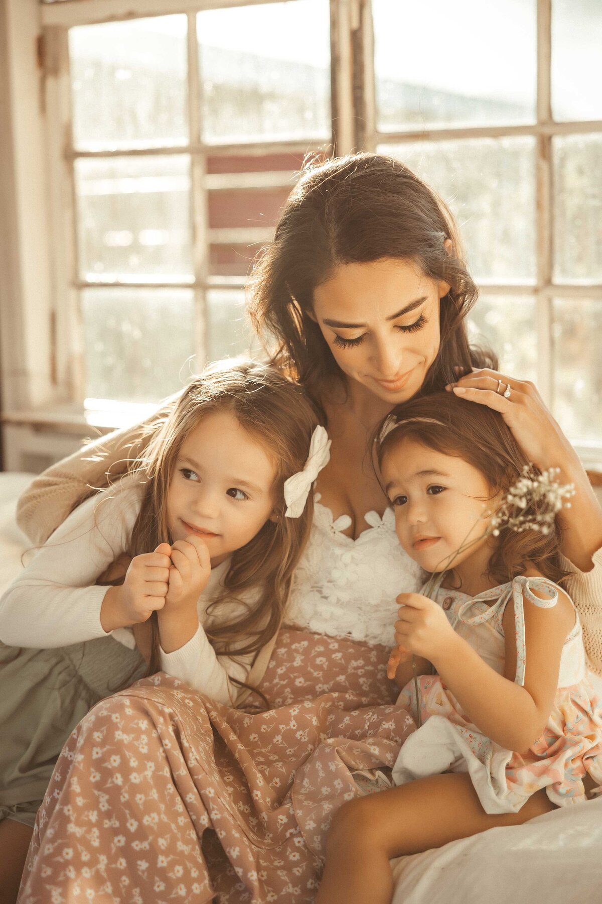 Woman wearing a white tops and ditsy flower skirt is hugging her two daughters who are holding a bunch of small flowers