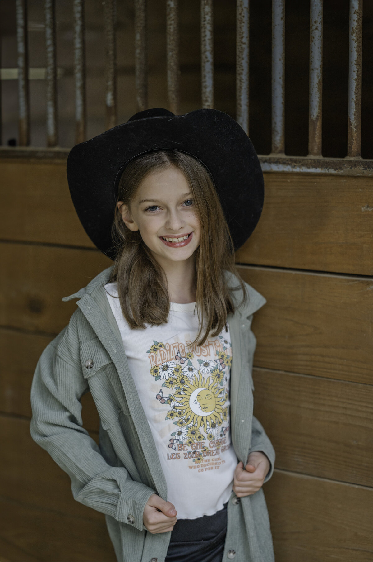 Little girl with brown hair wearing a cowboy hat smiles while in the horse stables