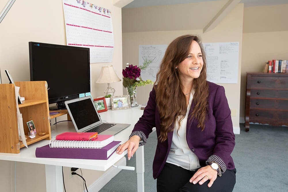 woman in a purple blazer seated at a desk looking to the right and smiling