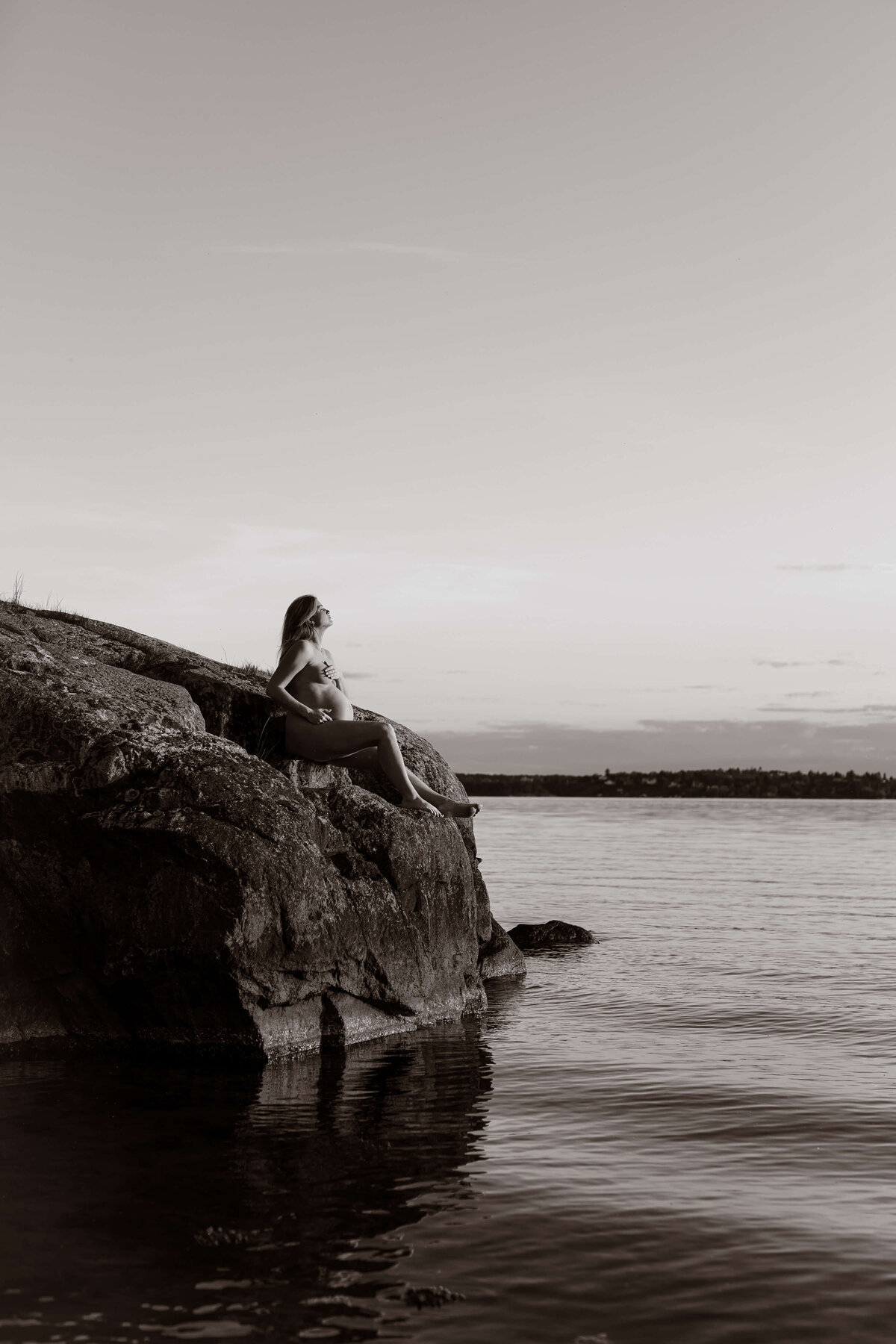 En svartvit bild på en gravid kvinna sittandes längre ifrån på ett berg ute vid vattnet under en gravidfotografering i Stockholm
