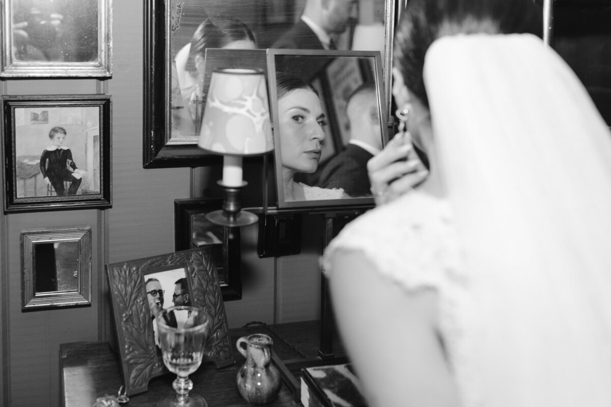 Bride looking at small wall mirror and fixing earrings while getting ready