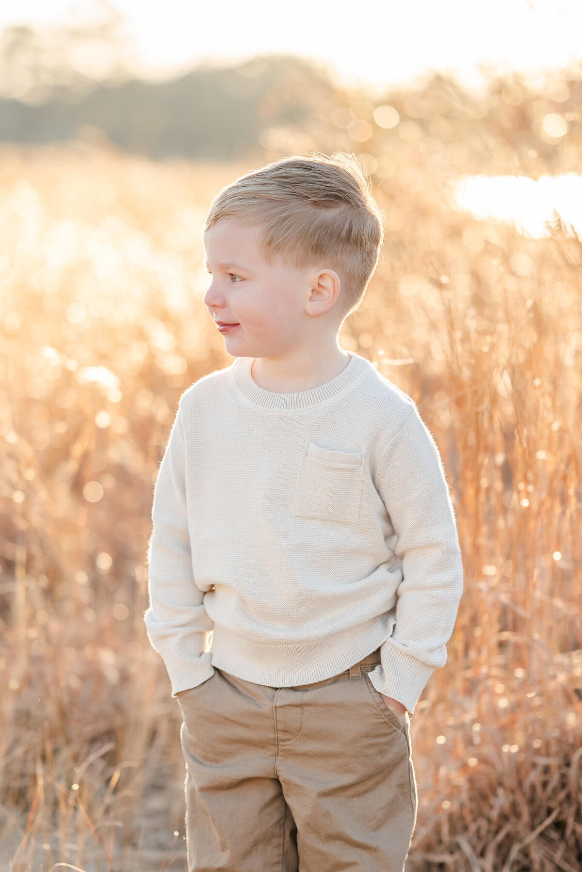 A young boy, wearing khakis and an off white top, puts his hands in his pockets and looks off to the side. He is standing in a field of brown grass with the sun shining behind him.