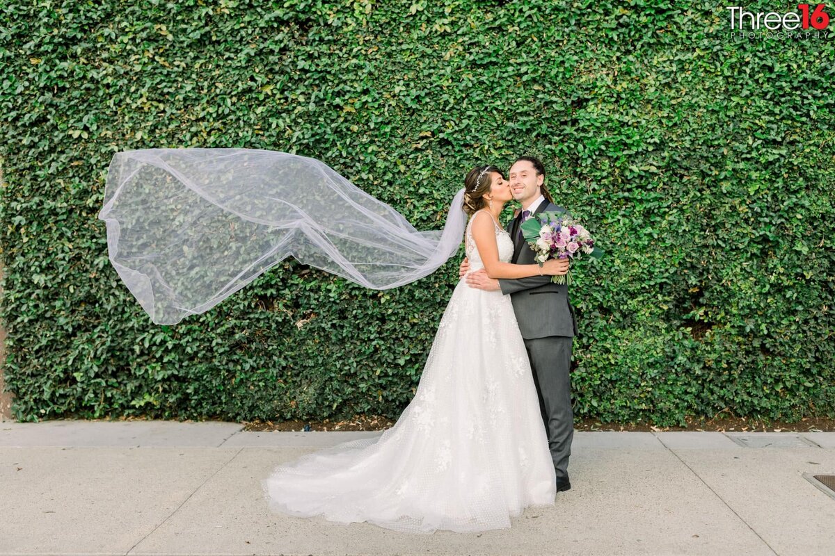 Bride kisses her Groom on the cheek while her veil flows in the wind