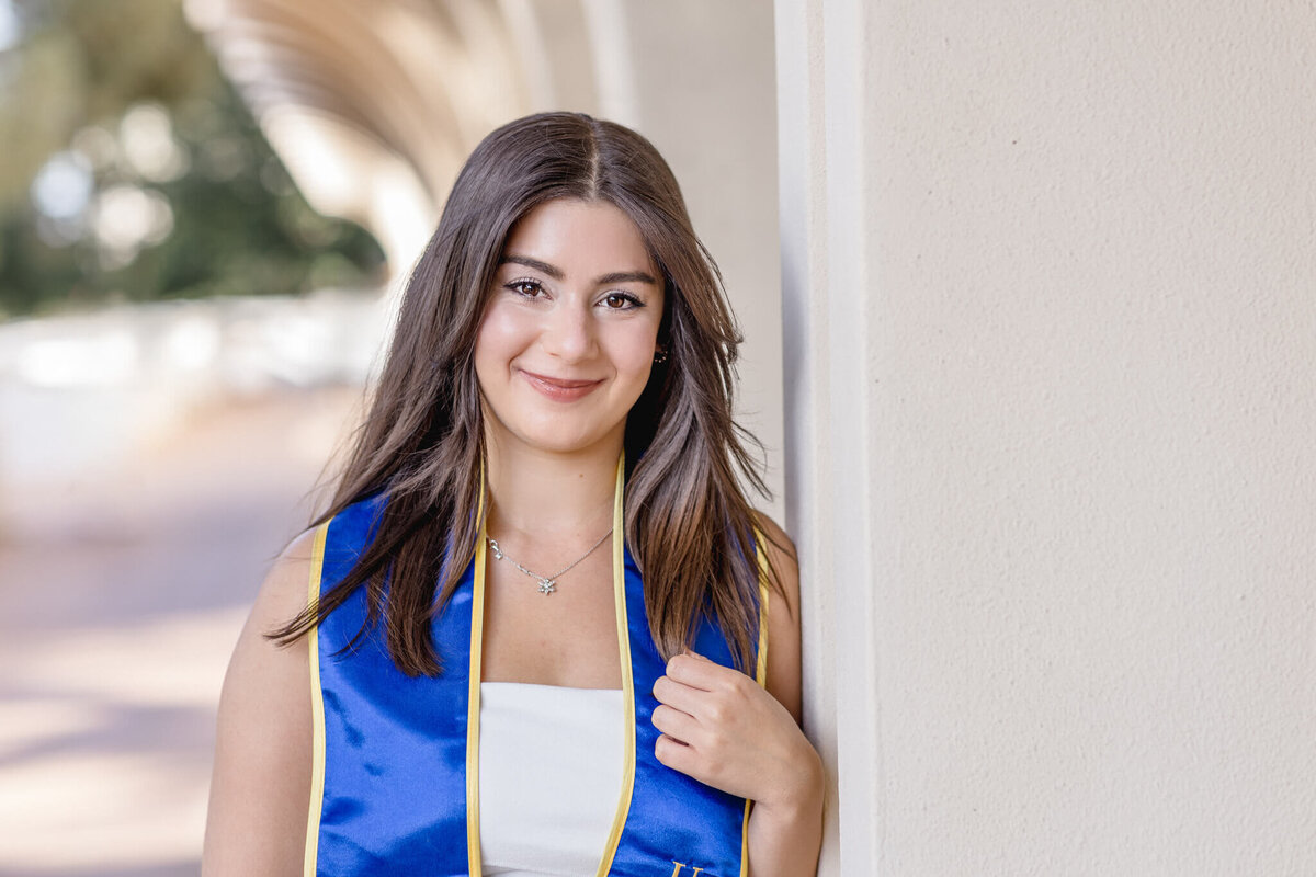 A young woman with long brown hair smiles while leaning against a light-colored wall. She is wearing a blue graduation stole over a white top. The background features an arched walkway with soft, natural lighting.