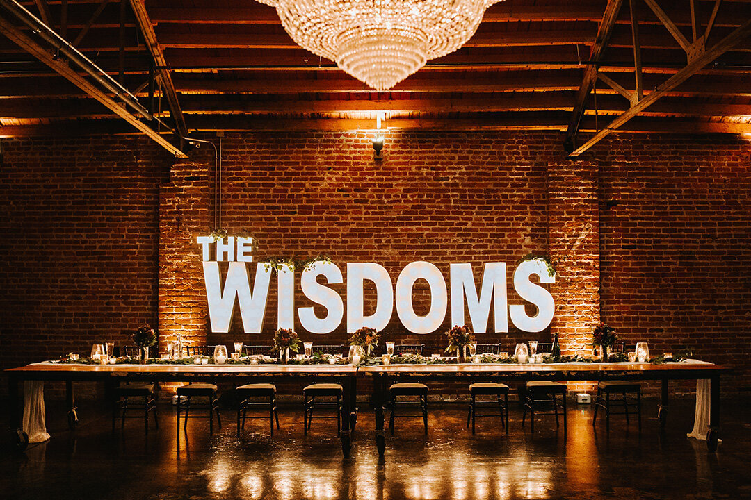 A lighted sign reads ‘The Wisdoms’  on a brick wall behind the main table at the wedding reception .