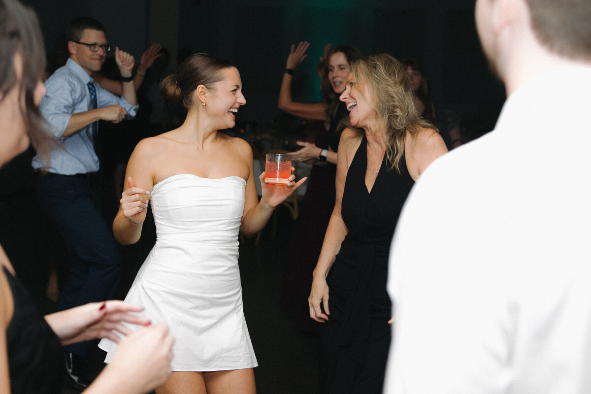 Bride in short, strapless reception dress holds her holds a cocktail dress with a pink beverage in it while dancing. She smiles over at a women who laughs and shrugs in her direction in a black floor length dress with a deep V neckline. Wedding guests dance behind them with their hands in the air. 