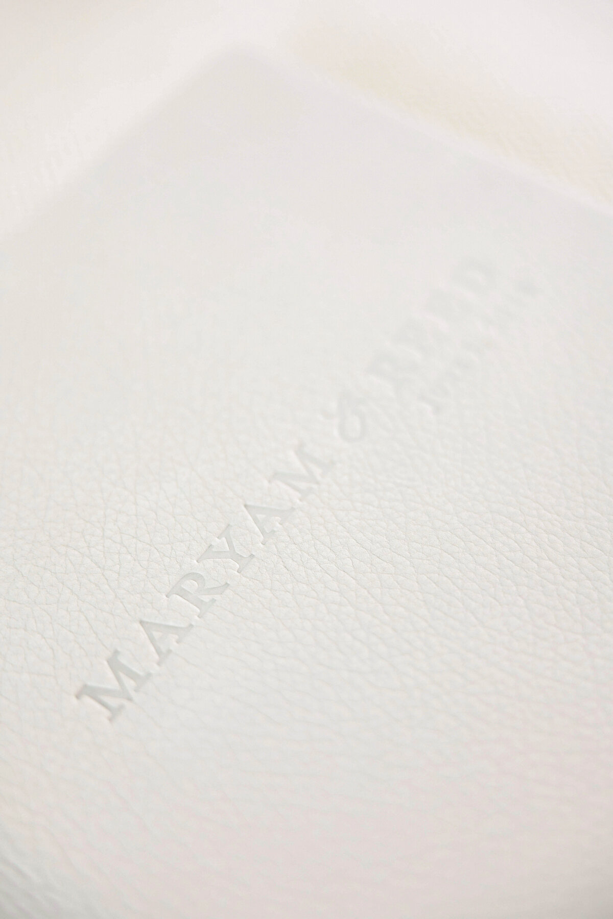 Close-up of a white textured surface with embossed text that reads MARYAM & READ. The focus highlights the leathery pattern and subtle shadows created by the embossing.