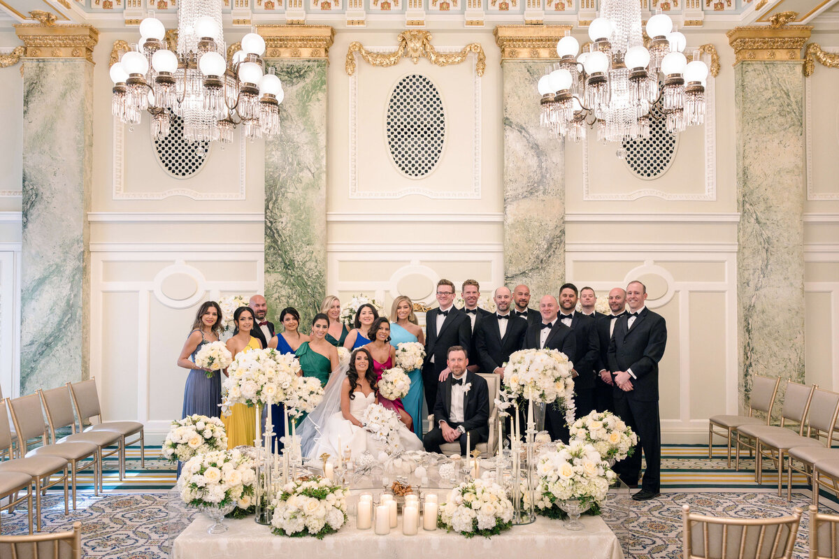 A formal wedding scene in an opulent room with chandeliers and marble walls. The couple is seated in front, surrounded by a large wedding party. The setting is elegantly decorated with white flowers and candles.