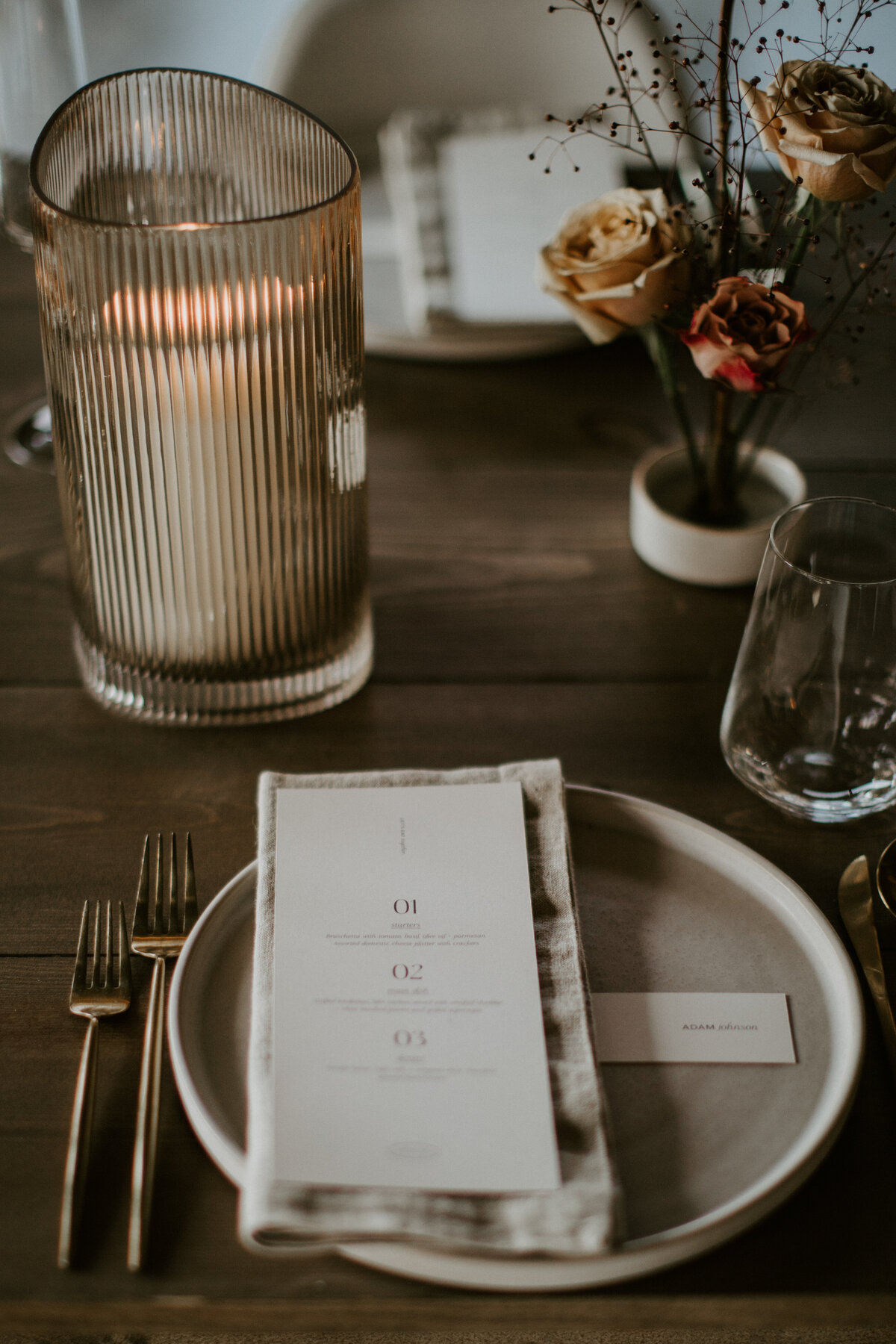 White dinner menu set on a white linen napkin and white plate atop a wooden table with gold silverware, flowers and a candle.