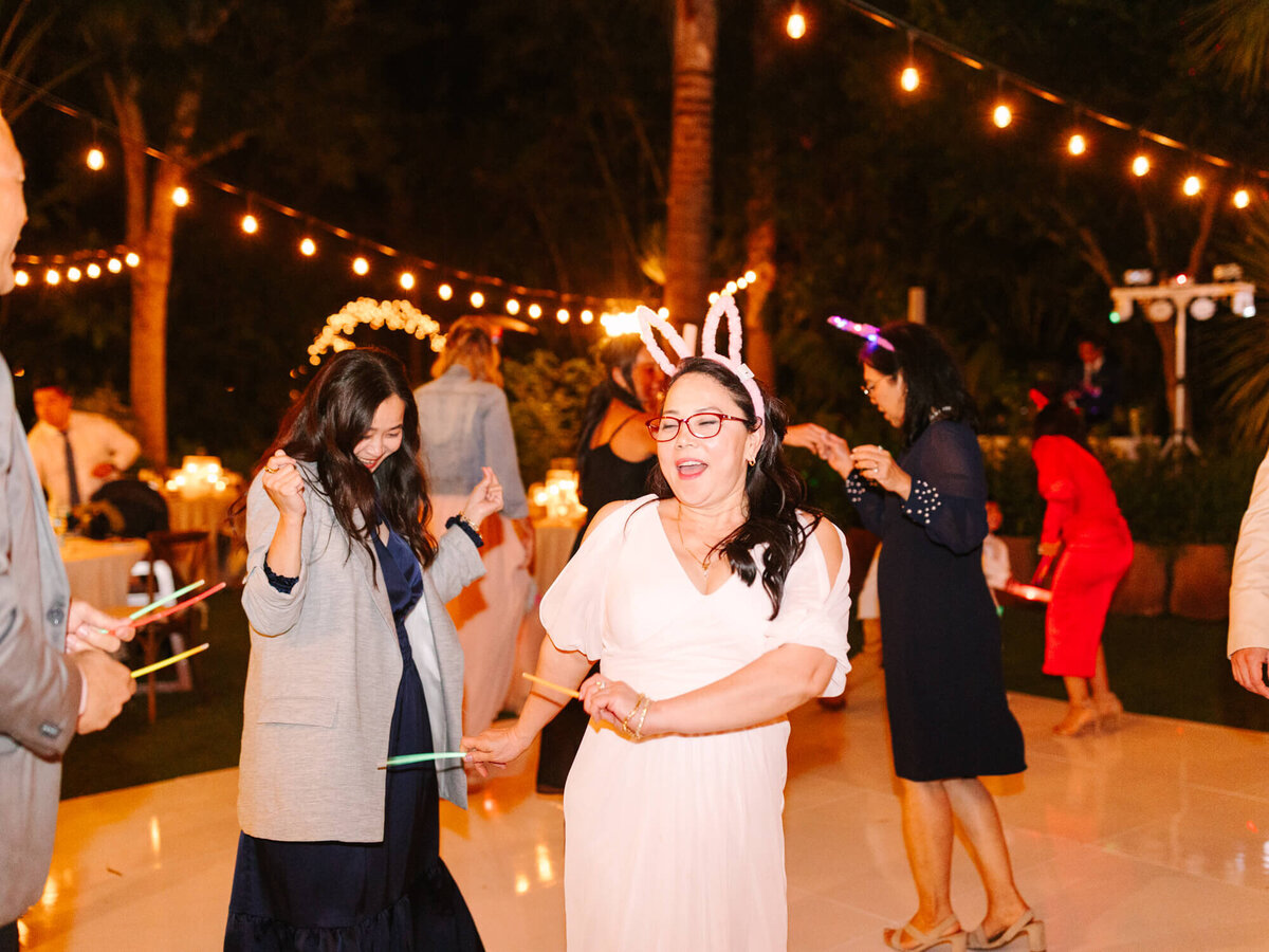 A festive reception with woman in a pink dress wearing bunny ears, dancing with friends under string lights.