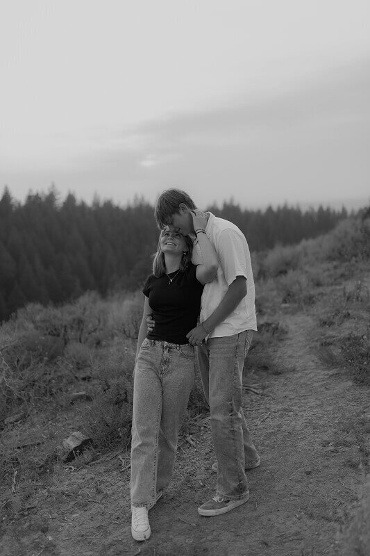 Black and white photo of a woman looking up at her boyfriend as he holds her from behind. Behind them are the trees and landscape of Rexburg, ID.