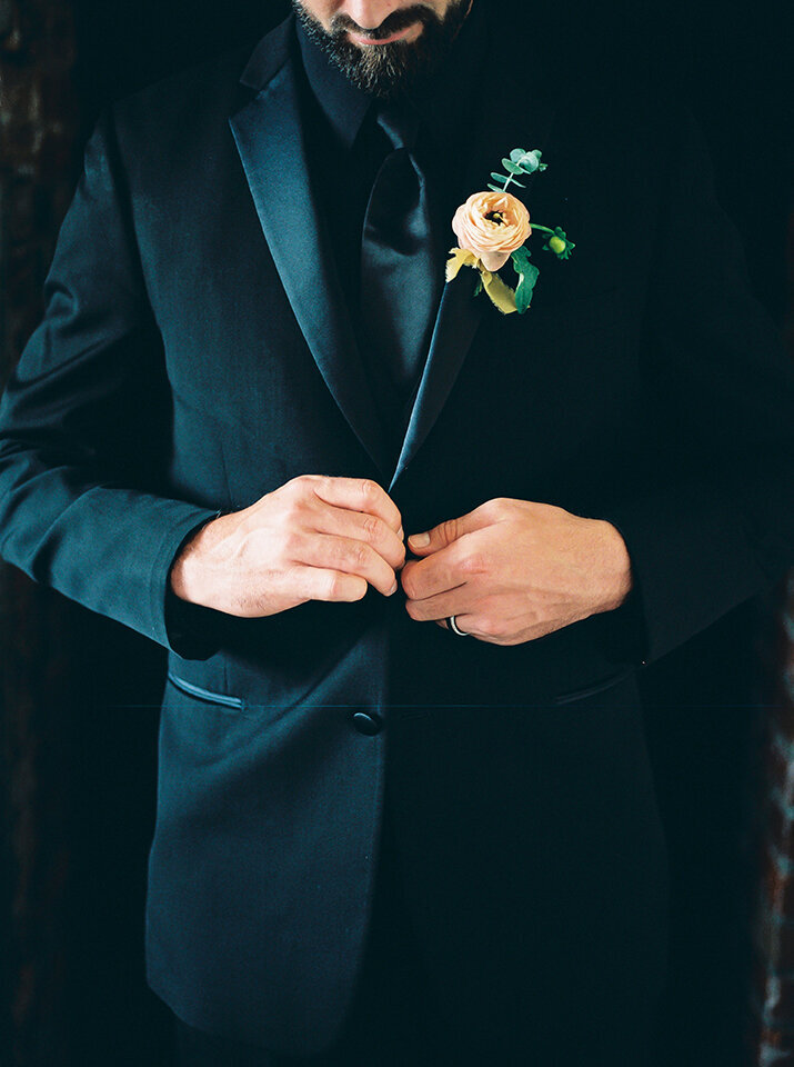 Close-up of groom wearing black tuxedo with ivory flower unbuttons his blazer.