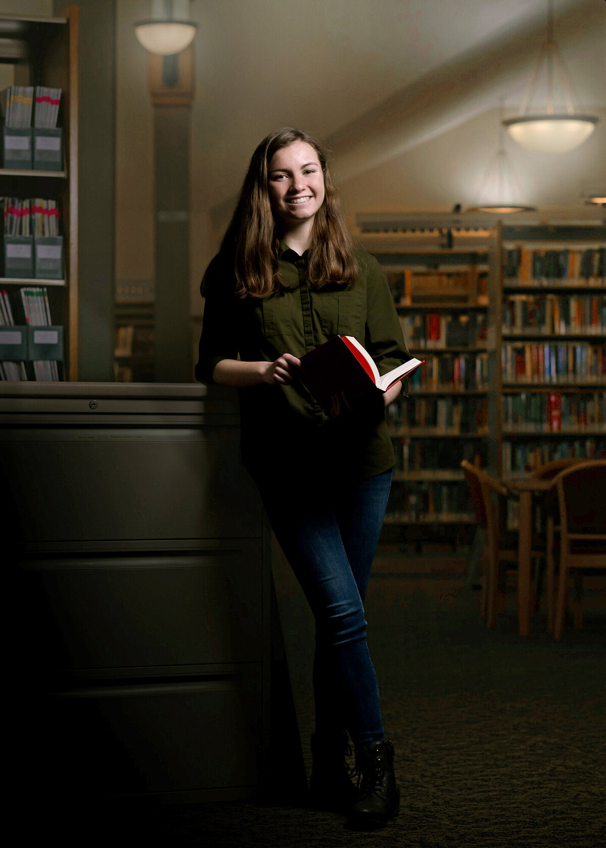 A high school senior stands in a library with a book in her hands in jeans and green shirt for a Pella Senior Photographer