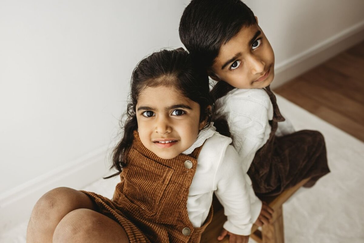 Two young siblings, a boy and a girl, look up at the camera. The girl wears a brown corduroy dress.