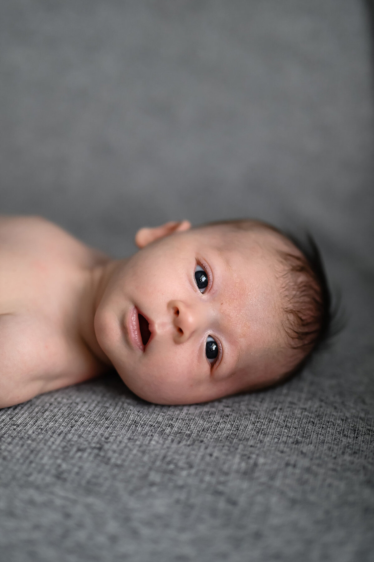 A newborn baby lays on a grey bed with eyes open
