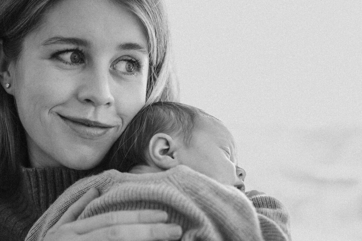 Mother holding her newborn over her shoulder, looking softly to the side while the baby sleeps against her chest.