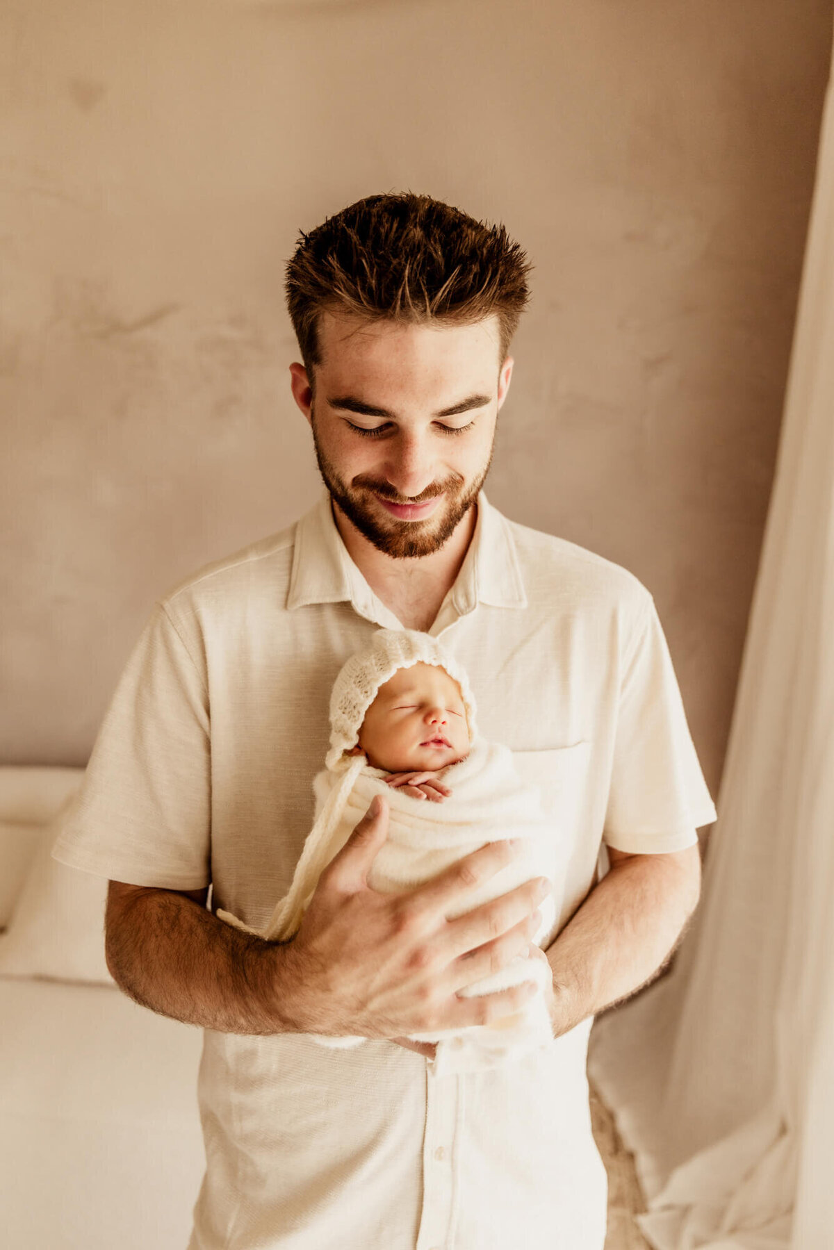 Father smiling down at his newborn daughter with a beige wall behind him.