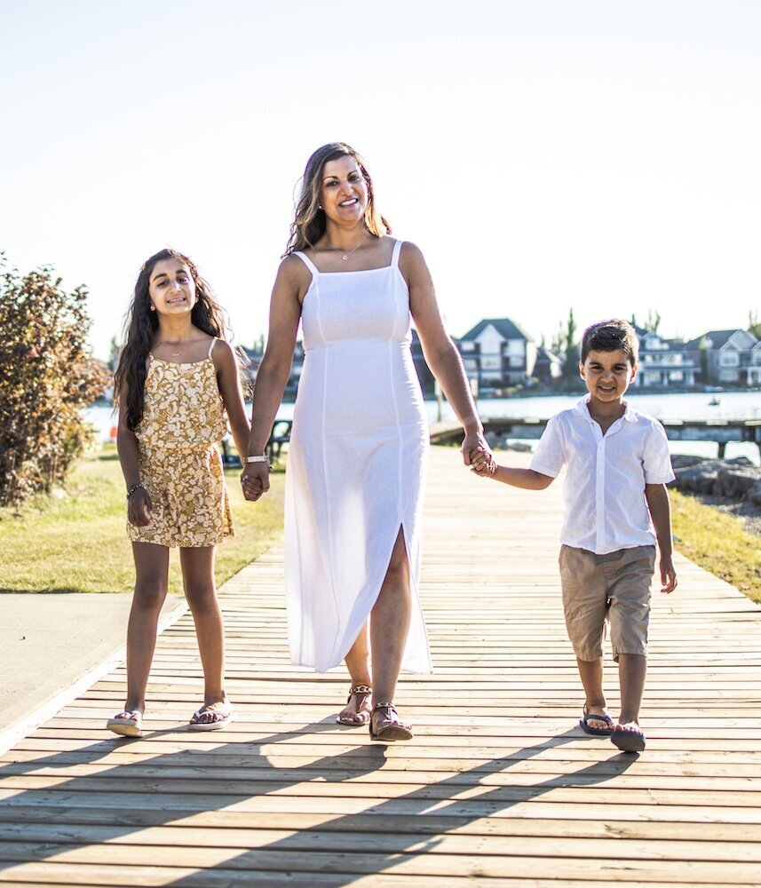 A mom in a white dress is walking near a lake holding her son and daughter's hands