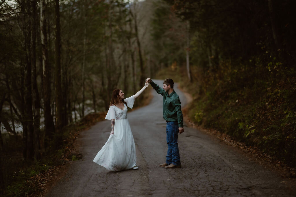 Bride and groom dancing on a quiet forest road near Greenbrier while eloping to Gatlinburg, with the groom twirling the bride in her white dress under the soft light of a wooded mountain path.

