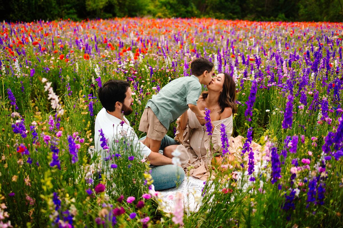 The professional maternity photographer captures a stunning family moment in a garden adorned with purple and pink flowers. The family, dressed in vintage outfits, is sitting on the grass, showcasing a warm and intimate scene filled with natural beauty and love.