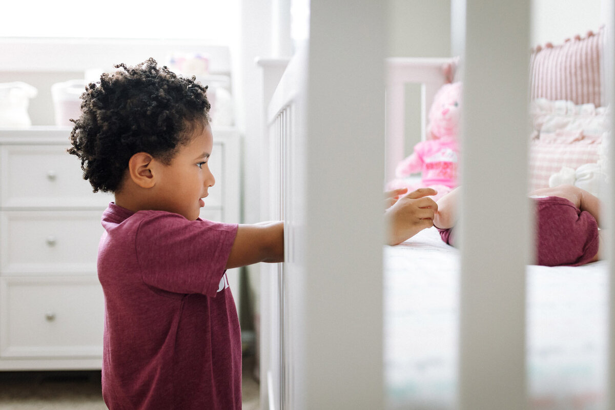 4-year-old boy reaching into crib to touch newborn sister's feet and wearing matching purple shirts