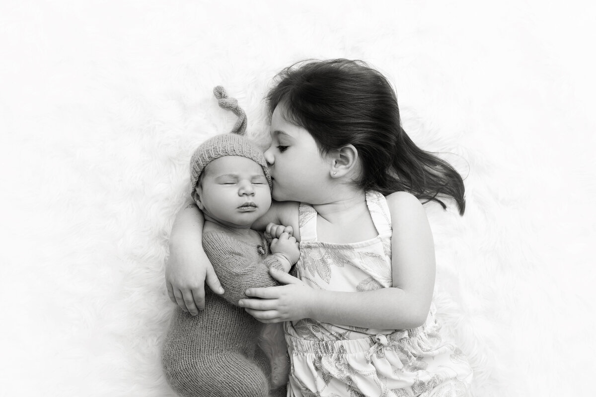 A toddler girl ina. dress lays on a bed kissing and cuddling with her sleeping newborn baby sibling as directed by a NJ Newborn Photography