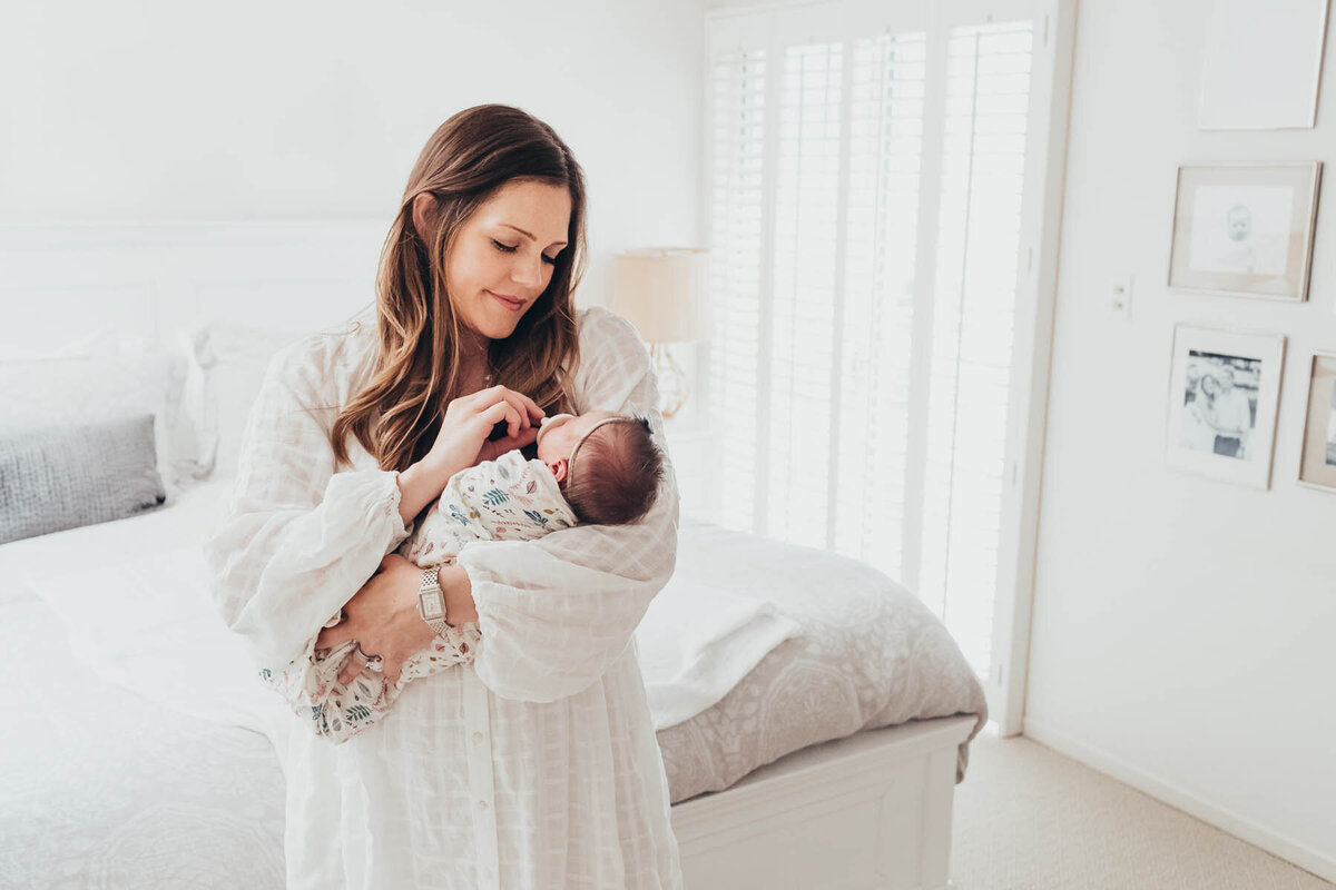 A beautiful mother holds her newborn baby in her arms while standing in her La Jolla home.