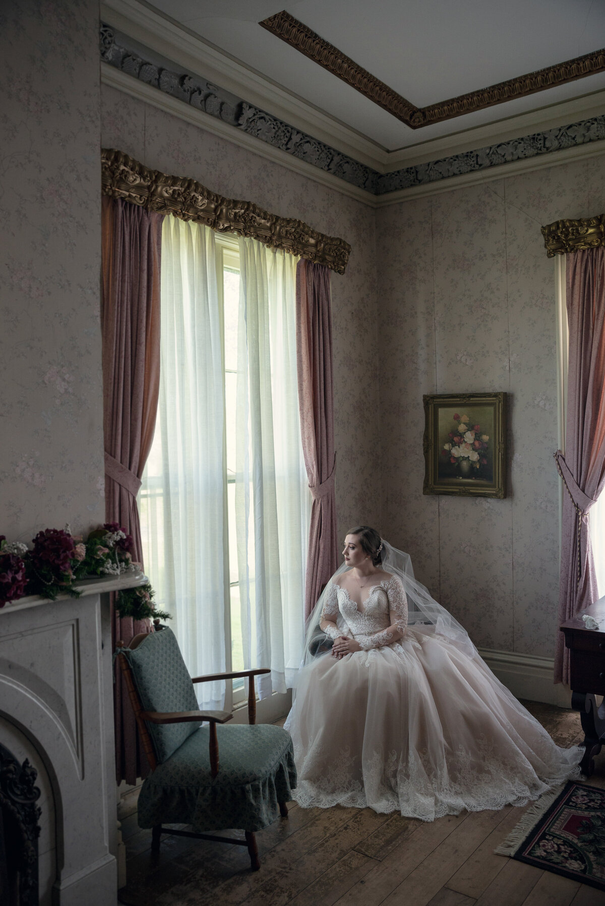 Bride sitting looking out window in front of a vintage painting.