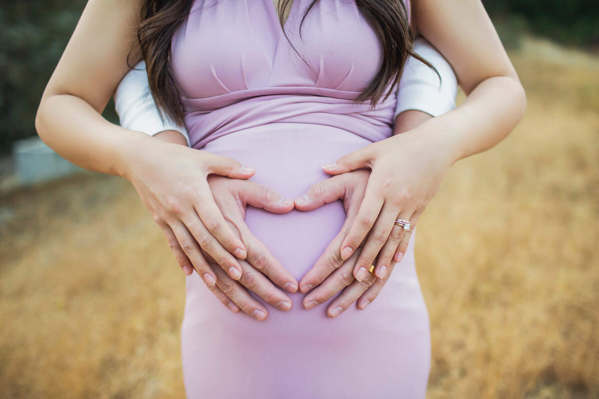 A close-up shot of a pregnant woman's belly, with her and her partner's hands forming a heart shape over her baby bump. She is wearing a form-fitting lavender dress, and her partner's arms are wrapped around her from behind, adding a sense of love and anticipation. The background is a golden field, creating a warm and intimate atmosphere.