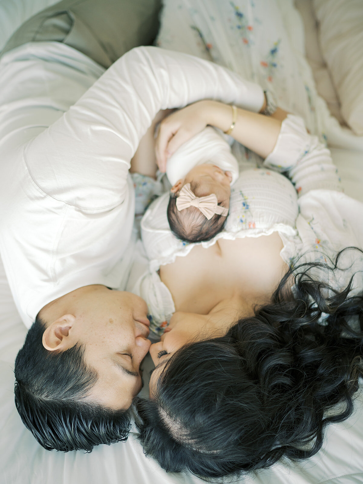 A cozy in-home lifestyle newborn session featuring parents snuggling their newborn while lying on their bed. The baby rests peacefully between them as they gaze lovingly at their little one. Soft natural light fills the room, creating a warm and intimate atmosphere that captures the joy and tenderness of this special moment.
