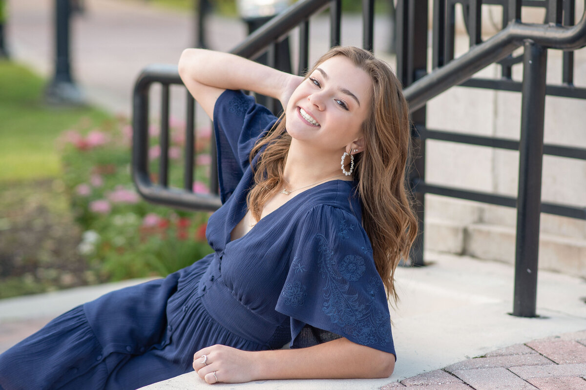 High school senior girl in blue dress lays on stairs with hand in hair smiling.