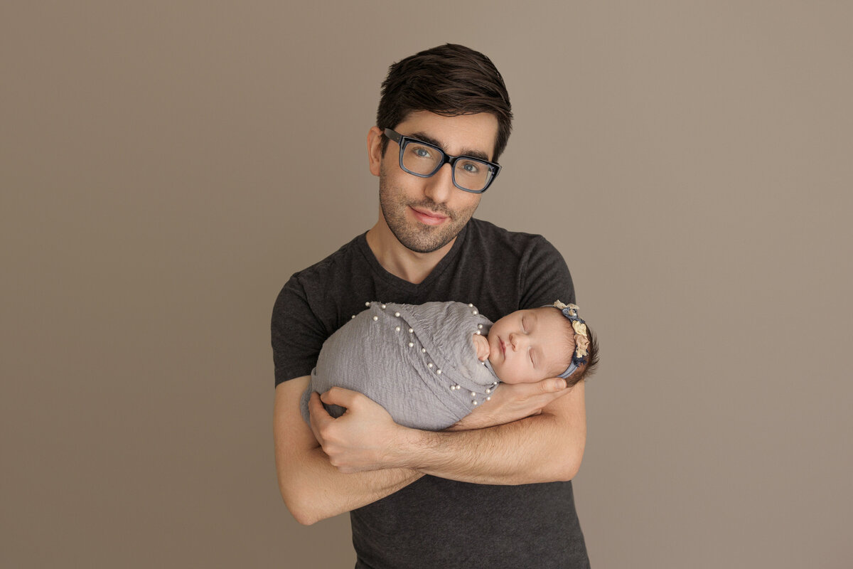 Father holding his sleeping newborn baby wrapped in a grey blanket, posing against a neutral beige background, father smiling softly.